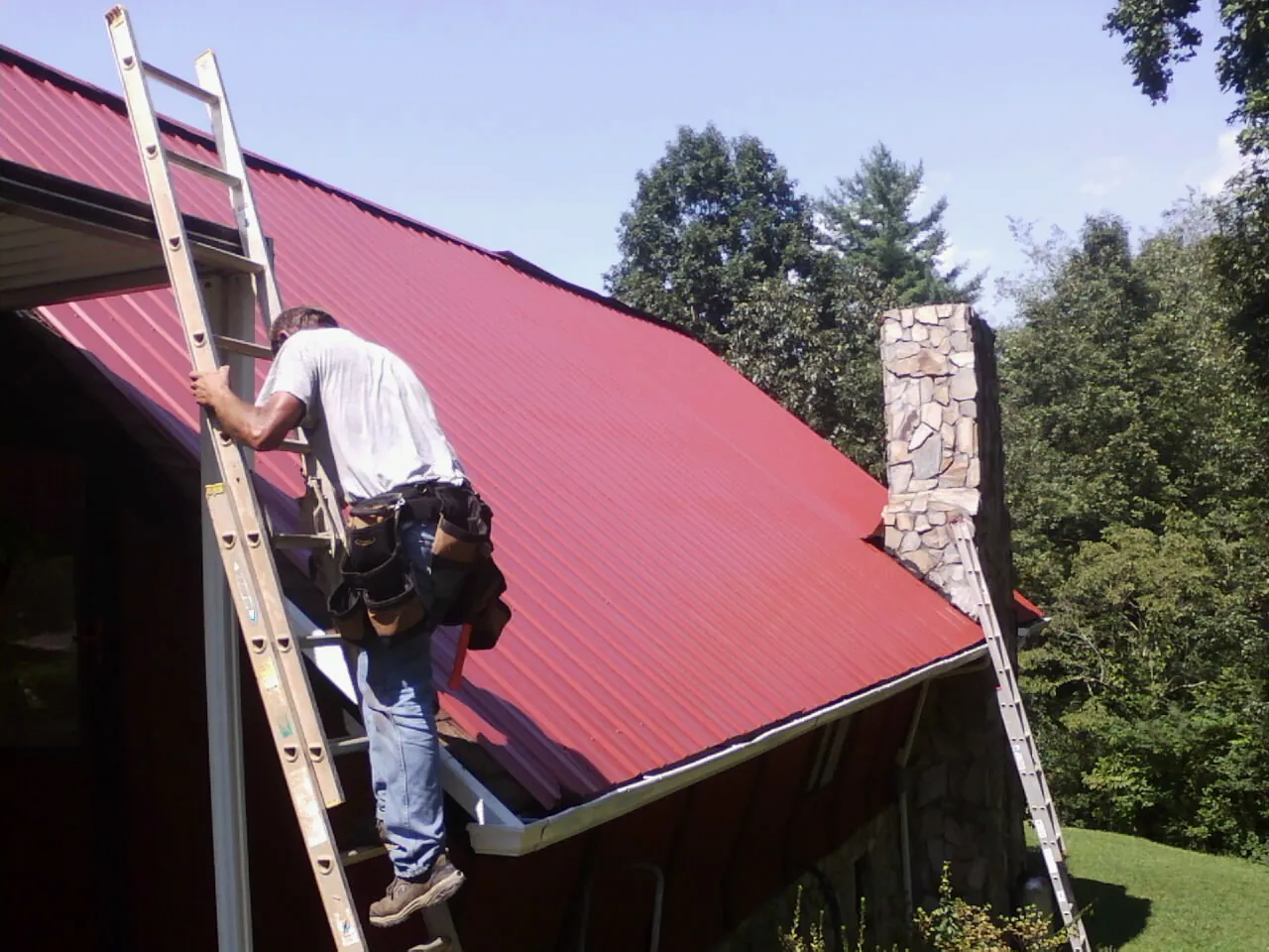 Roofer on a ladder beside a red metal roof and stone chimney — Soffit & Fascia Repair by Roofing Brothers in Bonlee